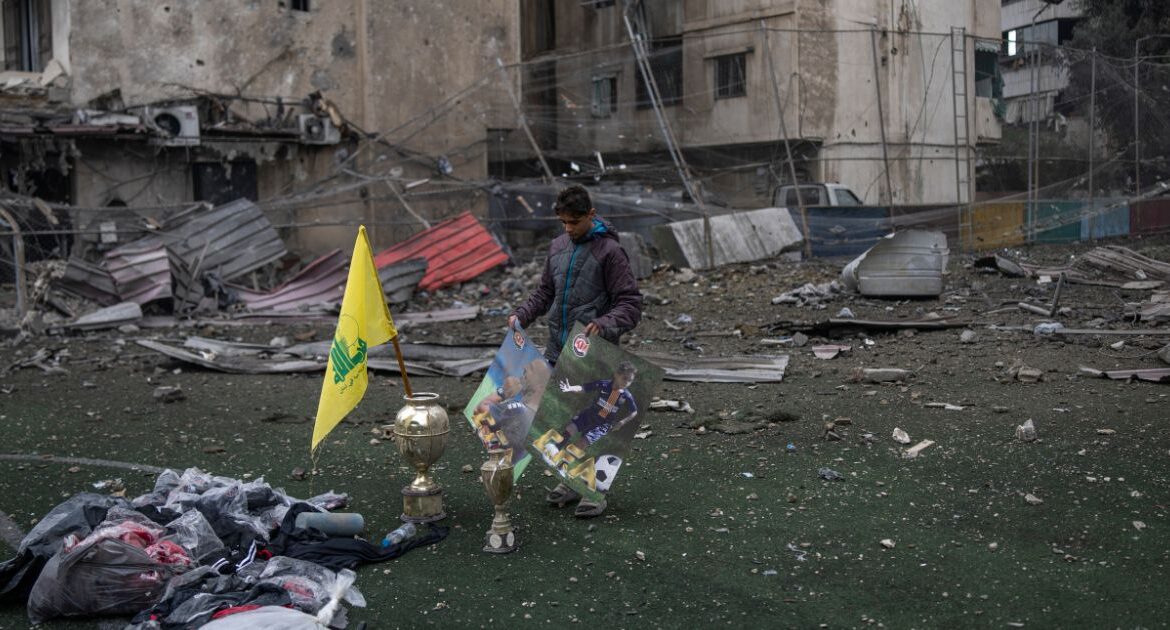 A young man salvages equipment and posters from a soccer field damaged by an airstrike as a Hezbollah flag sits in a trophy in Beirut, on Wednesday, Nov. 27, 2024. Thousands of people displaced by a year of fighting between Israel and Hezbollah packed roads leading to southern Lebanon on Wednesday as a U.S.-backed cease-fire began to take hold, bringing hope for an end to the deadliest war between the two sides in decades. (Diego Ibarra Sánchez/The New York Times)