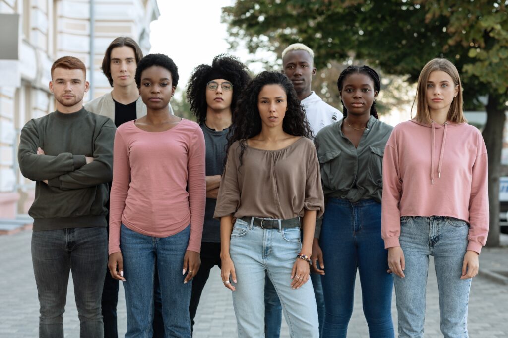 A group of young adults of different racial and ethnic backgrounds stand side by side outdoors, looking directly at the camera with serious expressions.