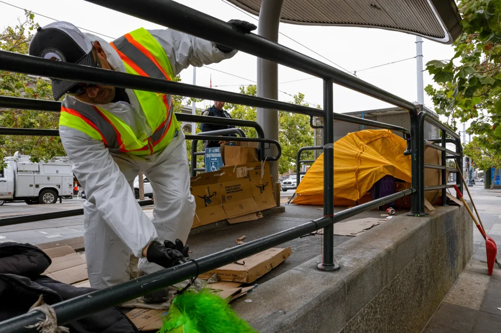 City worker in protective gear removes belongings near a yellow tent during a homeless encampment cleanup at an urban transit stop.